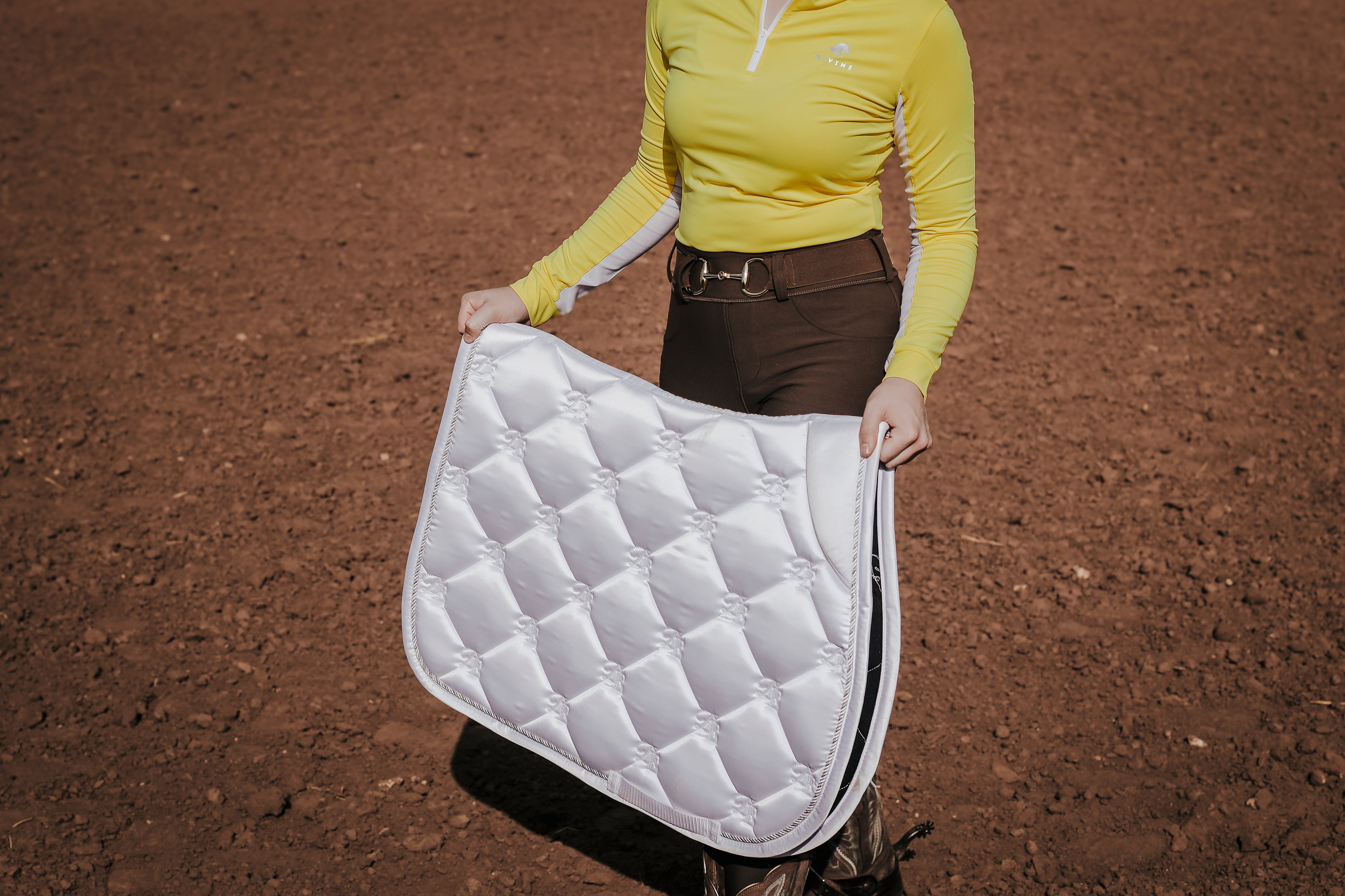 Finley Wright holding Divine Equestrian White Sovereign Saddle Pad, while wearing Yellow Sweet Lady Sun Shirt, Brown Comfort Fit Belt, and Mocha Comfort Grip Leggings